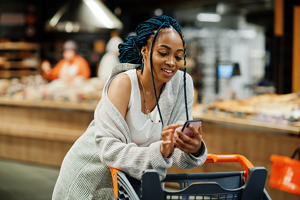 A woman smiling down at her smart phone leaning on a shopping cart in a store
