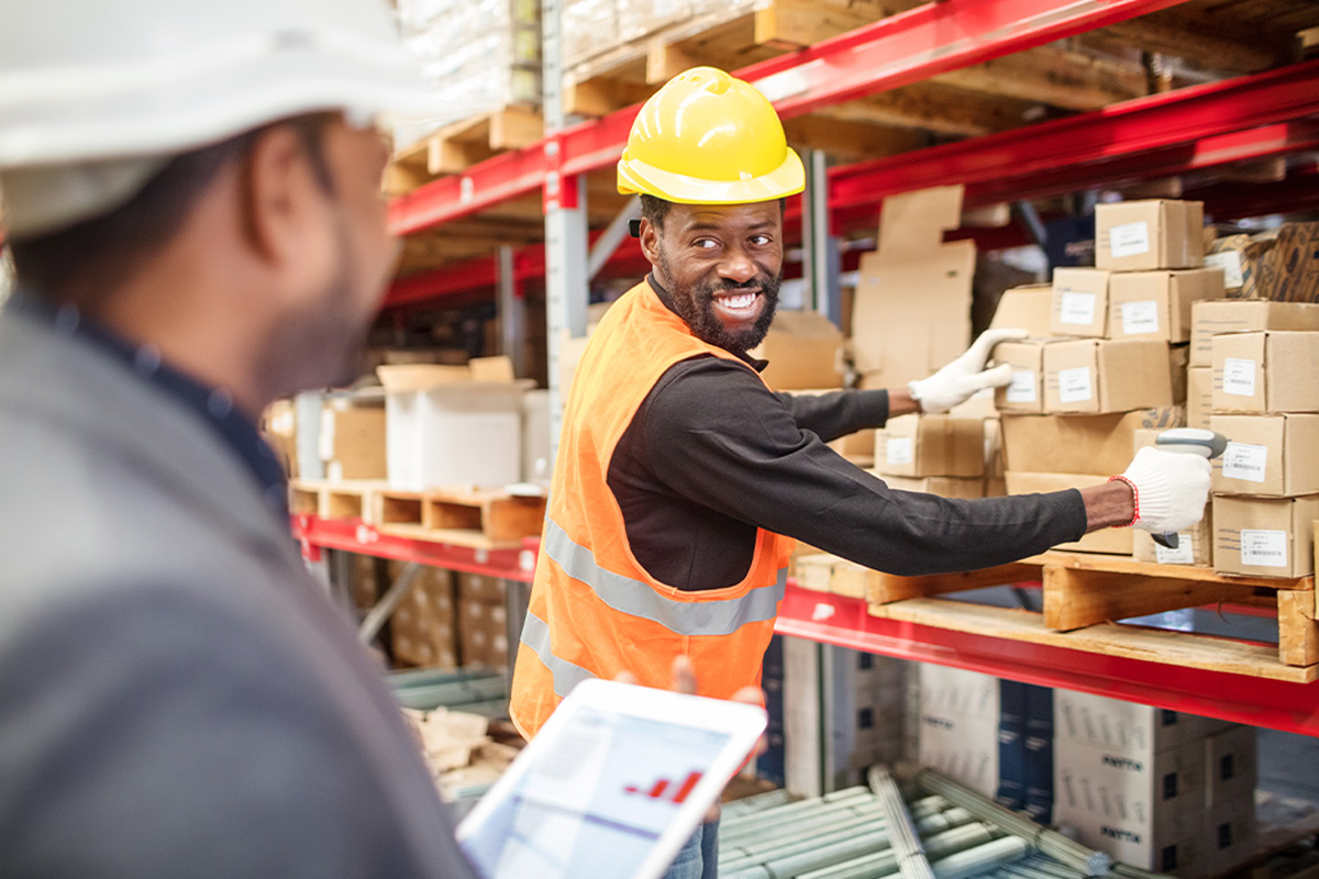 A man in a hard hat and orange west smiling at another man in a warehouse scanning a code on a cardboard box