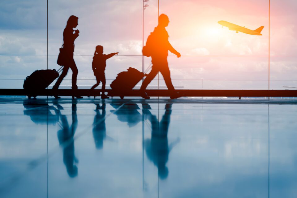 a three member family walking through the airport with luggage as the sunsets in the distance and a plane taking off